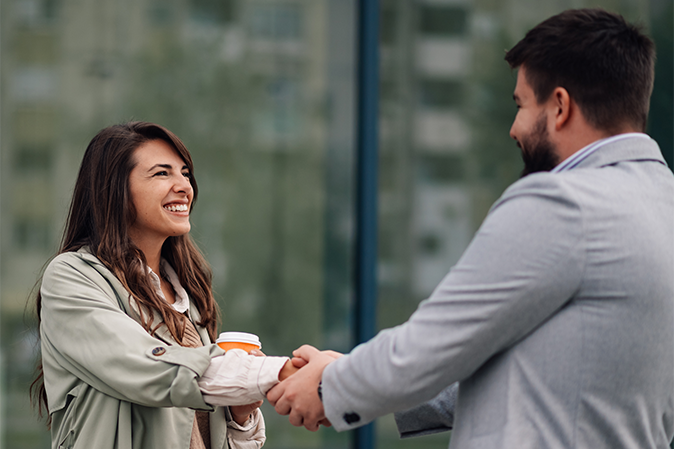Two business people are shaking hands in front of an office building while one of them is holding a cup of coffee and smiling